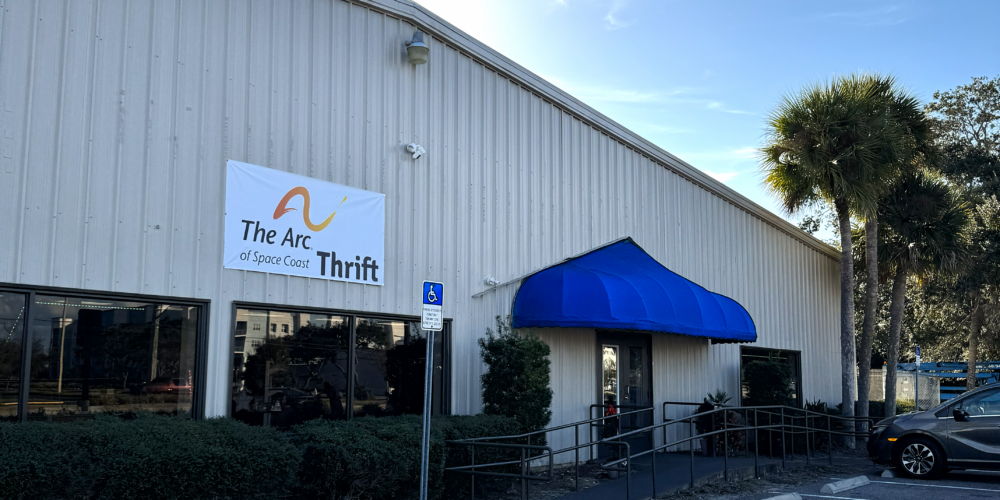 Image of the store front. The building is tan in color with a ramp leading to the entrance, which is covered by a blue awning. The sun is just behind the building, and a palm tree is lit up to the right.